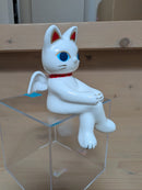White cat-like figurine with red accents sitting on a clear stand against a wooden background.