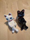 Two ceramic cat figurines, one white and one black, on a wooden floor.